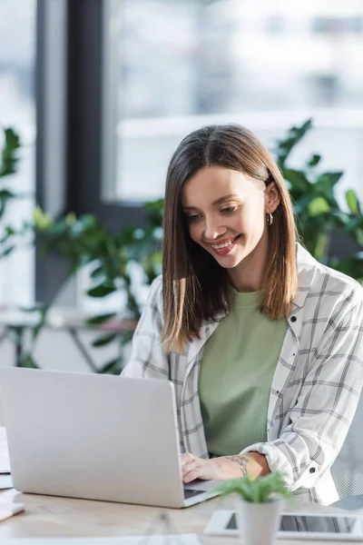 Fröhliche Geschäftsfrau mit Laptop in der Nähe von verschwommenem digitalem Tablet im Büro — Stockfoto