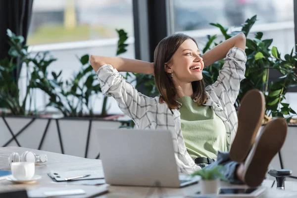 Femme d'affaires joyeuse assise près d'un ordinateur portable flou, café et écouteurs dans le bureau — Photo de stock