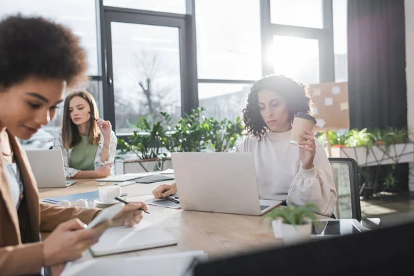 Empresaria musulmana sosteniendo café para acercarse a colegas interracial y dispositivos en la oficina - foto de stock