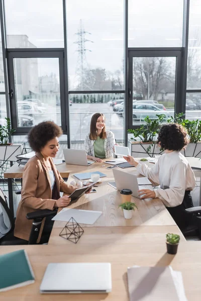 Positive interracial businesswomen talking near devices and papers on table in office — Stock Photo