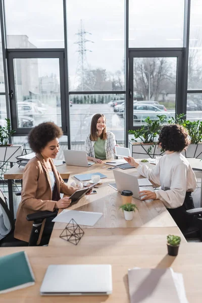 Positive interracial businesswomen talking near devices and papers on table in office — стоковое фото