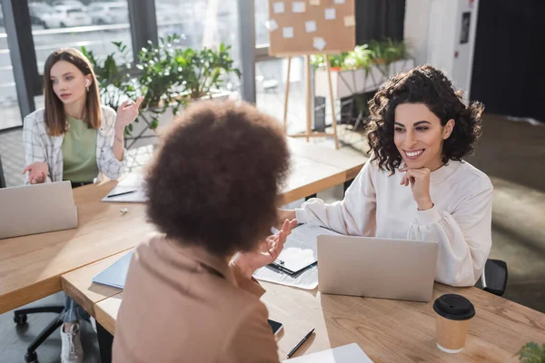 Alegre musulmana mujer de negocios mirando borrosa africana americana colega cerca de dispositivos y café en la oficina - foto de stock