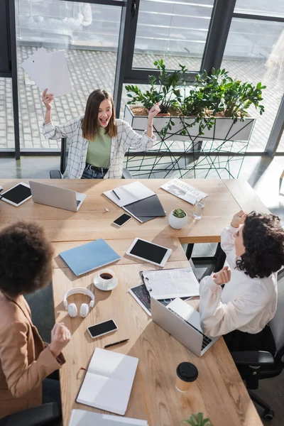 Overhead view of cheerful multiethnic businesswomen working near devices and papers on table — Stock Photo