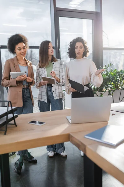 Arabian businesswoman holding documents and talking to interracial colleagues near blurred devices in office — Stock Photo