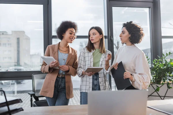 Lächelnde afrikanisch-amerikanische Geschäftsfrau im Gespräch mit multiethnischen Kollegen mit Papieren in der Nähe eines verschwommenen Laptops im Büro — Stockfoto