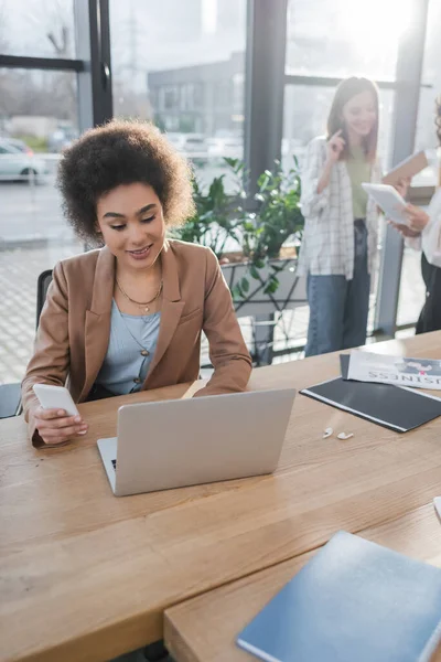 Alegre mujer de negocios afroamericana usando dispositivos cerca de auriculares y documentos en la mesa en la oficina - foto de stock