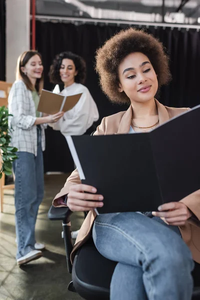 Cheerful african american businesswoman looking at paper folder near blurred interracial colleagues in office — стоковое фото