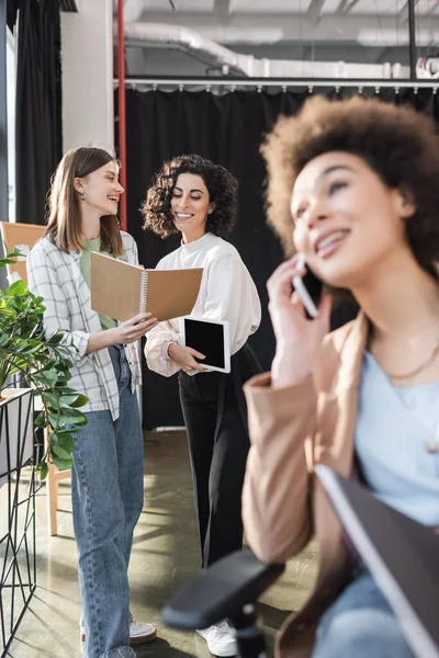 Positive multiethnic businesswomen looking at notebook while talking near blurred african american colleague talking on smartphone in office — Foto stock