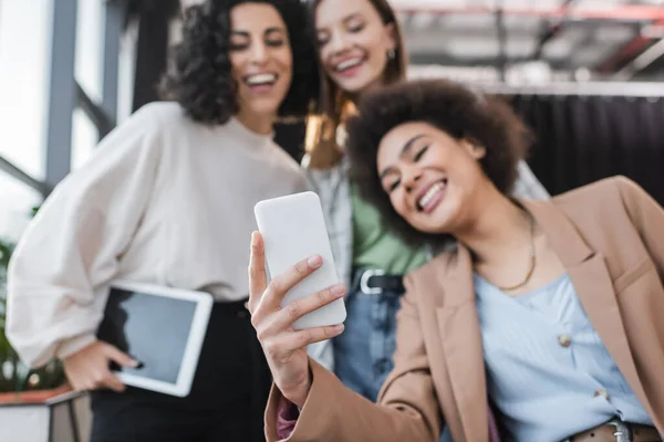 Empresarias multiétnicas borrosas sonriendo mientras se toman selfie en el teléfono inteligente en la oficina - foto de stock