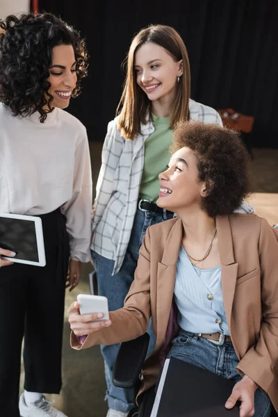 Alegre mujer de negocios afroamericana sosteniendo teléfono celular cerca de colegas multiétnicos en el cargo - foto de stock