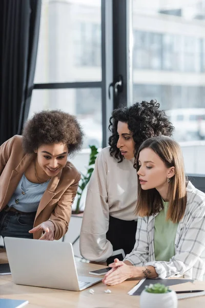 Alegre mujer de negocios afroamericana señalando a la computadora portátil cerca de colegas interracial en la oficina - foto de stock