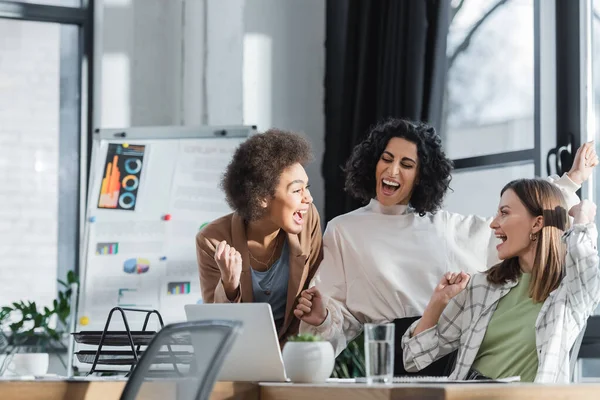 Emocionadas empresarias multiétnicas mirándose entre sí cerca de la computadora portátil en la oficina - foto de stock