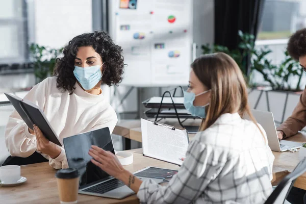 Donna d'affari musulmana in maschera medica che tiene quaderno vicino al collega con appunti e laptop in ufficio — Foto stock