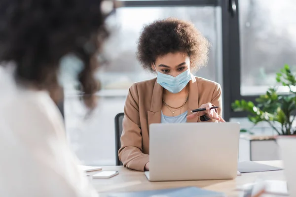 Donna d'affari afroamericana in maschera medica che tiene la penna vicino ai dispositivi e collega sfocato in ufficio — Foto stock