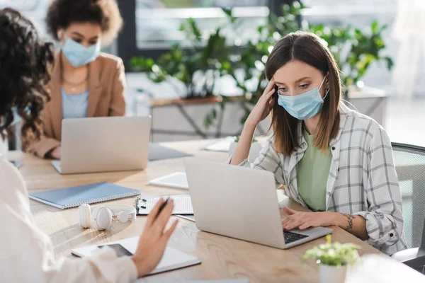 Businesswoman in medical mask using laptop near blurred interracial colleagues in office — стоковое фото