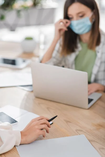 Donna d'affari che tiene la penna vicino al notebook, al cellulare e al collega sfocato in carica — Foto stock