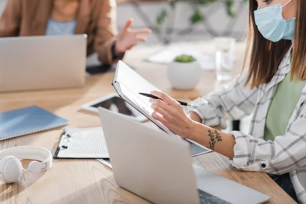 Vista cortada de empresária em máscara médica segurando notebook perto de laptop e colega turvo no escritório — Fotografia de Stock