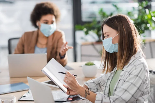 Businesswoman in medical mask holding notebook near blurred laptop and african american colleague in office — стоковое фото