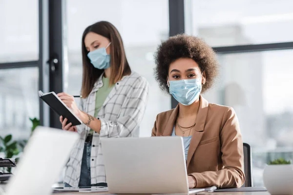 African american businesswoman in medical mask looking at camera near laptop and colleague in office — стокове фото