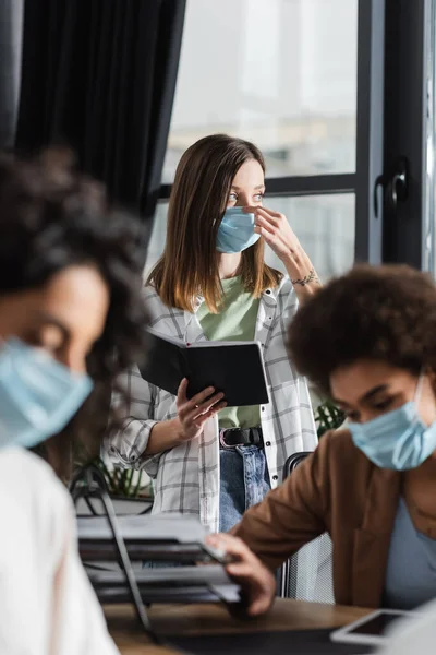 Joven empresaria sosteniendo un cuaderno y usando máscara médica cerca de colegas multiétnicos borrosos en la oficina - foto de stock