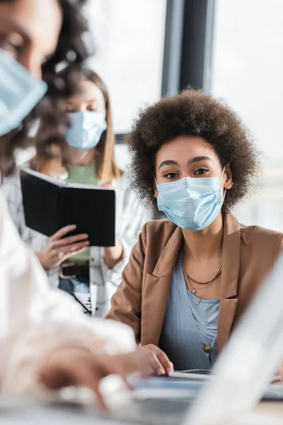 Afro americana empresaria en médico máscara mirando cámara cerca borrosa interracial colegas en oficina - foto de stock