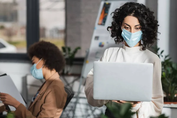 Donna d'affari musulmana in maschera medica utilizzando laptop vicino sfocato collega afro-americano in carica — Foto stock