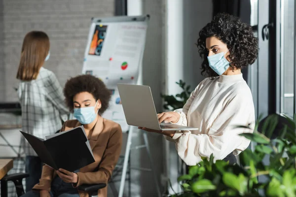 Arabo donna d'affari in maschera medica utilizzando laptop vicino pianta e colleghi interrazziali in ufficio — Foto stock