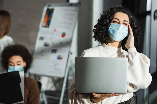 Muslim businesswoman in medical mask holding laptop while suffering from headache in office — стоковое фото