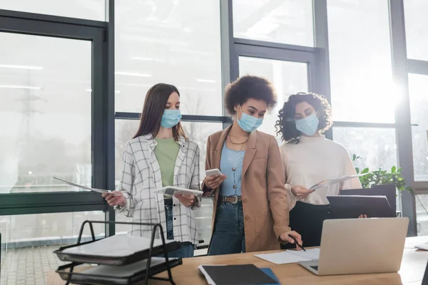 Donne d'affari multietniche in maschere mediche che detengono dispositivi e lavorano con documenti in carica — Foto stock