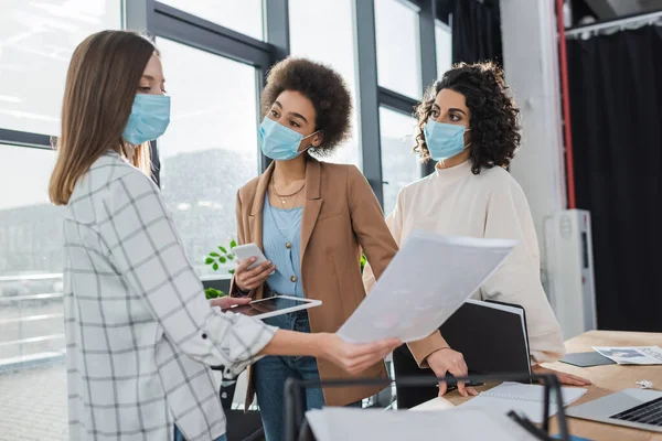 Multiethnic businesswomen in medical masks looking at colleague with digital tablet and document in office — Fotografia de Stock