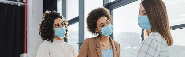 Multikulturelle Geschäftsfrauen in medizinischen Masken blicken auf Kollegin im Büro, Transparent — Stockfoto
