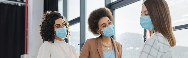 Multicultural businesswomen in medical masks looking at colleague in office, banner — стоковое фото