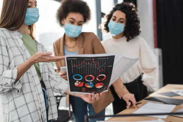 Empresaria en máscara médica apuntando al papel con cartas cerca de colegas multiétnicos borrosos en la oficina - foto de stock