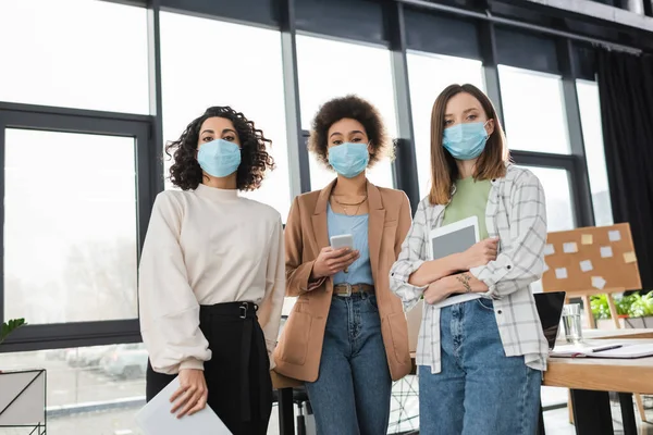 Empresarias interraciales en máscaras médicas sosteniendo dispositivos y papeles en la oficina - foto de stock