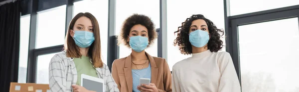 Multicultural businesswomen in medical masks holding gadgets in office, banner — Stock Photo