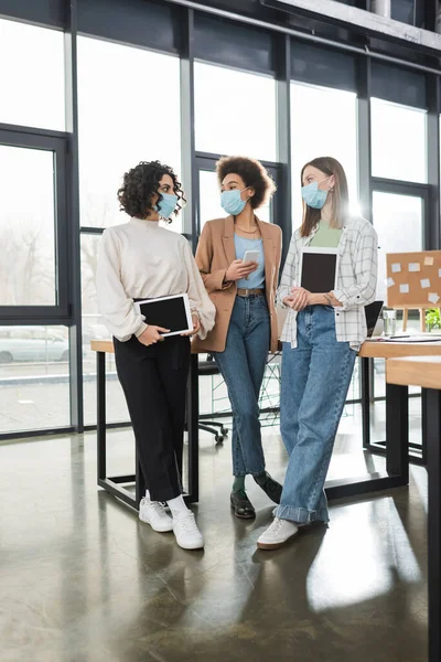 Multiethnic businesswomen in medical masks holding devices while talking in office — Stock Photo