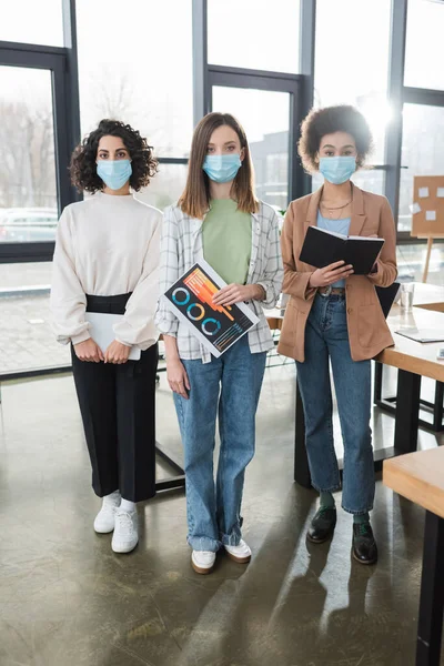 Multicultural businesswomen in protective masks holding digital tablet, paper and notebook in office - foto de stock