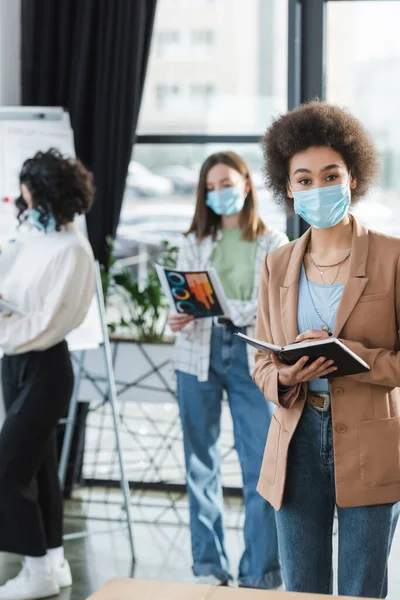 Empresaria afroamericana en máscara médica sosteniendo cuaderno mientras trabaja en la oficina - foto de stock