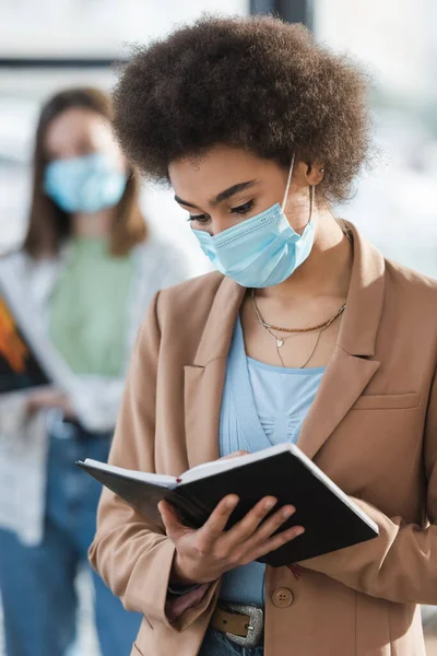 African american businesswoman in medical mask holding notebook in office — стокове фото