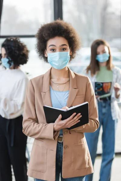 Young african american businesswoman in protective mask holding notebook and looking at camera in office — стокове фото