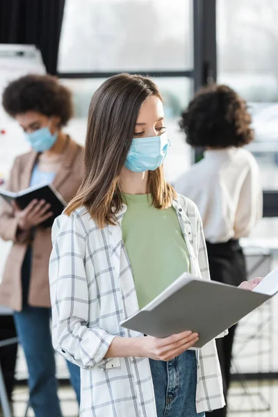 Empresaria con máscara médica sosteniendo una carpeta de papel borrosa cerca de colegas interracial borrosos en la oficina - foto de stock
