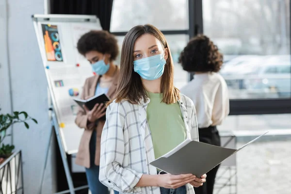 Joven mujer de negocios con máscara médica sosteniendo una carpeta de papel cerca de colegas multiétnicos borrosos - foto de stock