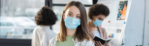 Businesswoman in protective mask looking at camera near blurred multiethnic colleagues in office, banner — Photo de stock
