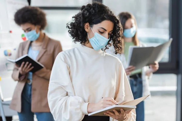 Mulher de negócios árabe em máscara médica escrevendo no caderno perto de colegas turvos no escritório — Fotografia de Stock