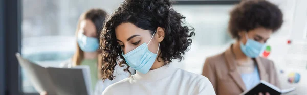 Muslim businesswoman in medical mask standing near blurred colleagues in office, banner - foto de stock