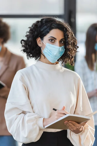 Mujer de negocios árabe en máscara protectora sosteniendo pluma y cuaderno mientras mira a la cámara en la oficina - foto de stock