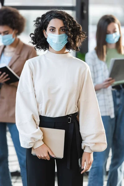 Musulmana mujer de negocios en máscara médica sosteniendo portátil y mirando a la cámara en la oficina - foto de stock