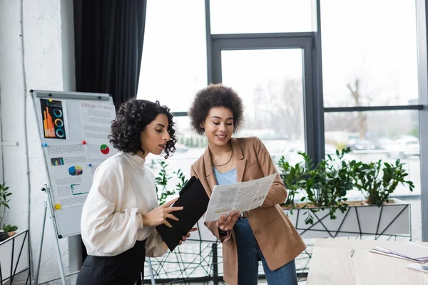 Positiva mujer de negocios afroamericana sosteniendo periódico cerca de colega musulmán en el cargo - foto de stock