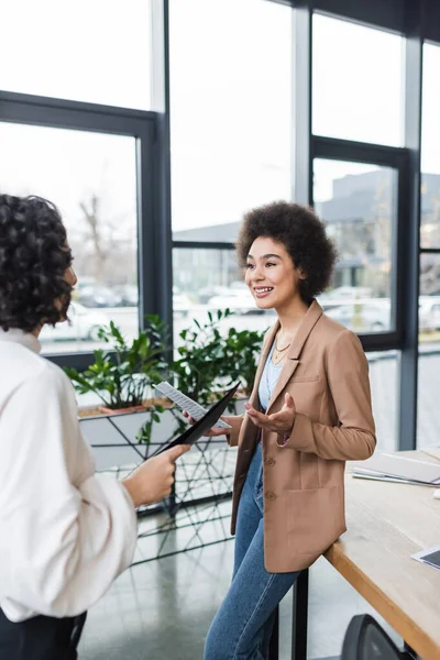 Alegre mujer de negocios afroamericana sosteniendo periódico cerca borrosa colega musulmán en la oficina - foto de stock