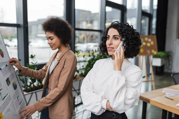 Young arabian businesswoman talking on smartphone near blurred african american colleague and flip chart in office — Stock Photo
